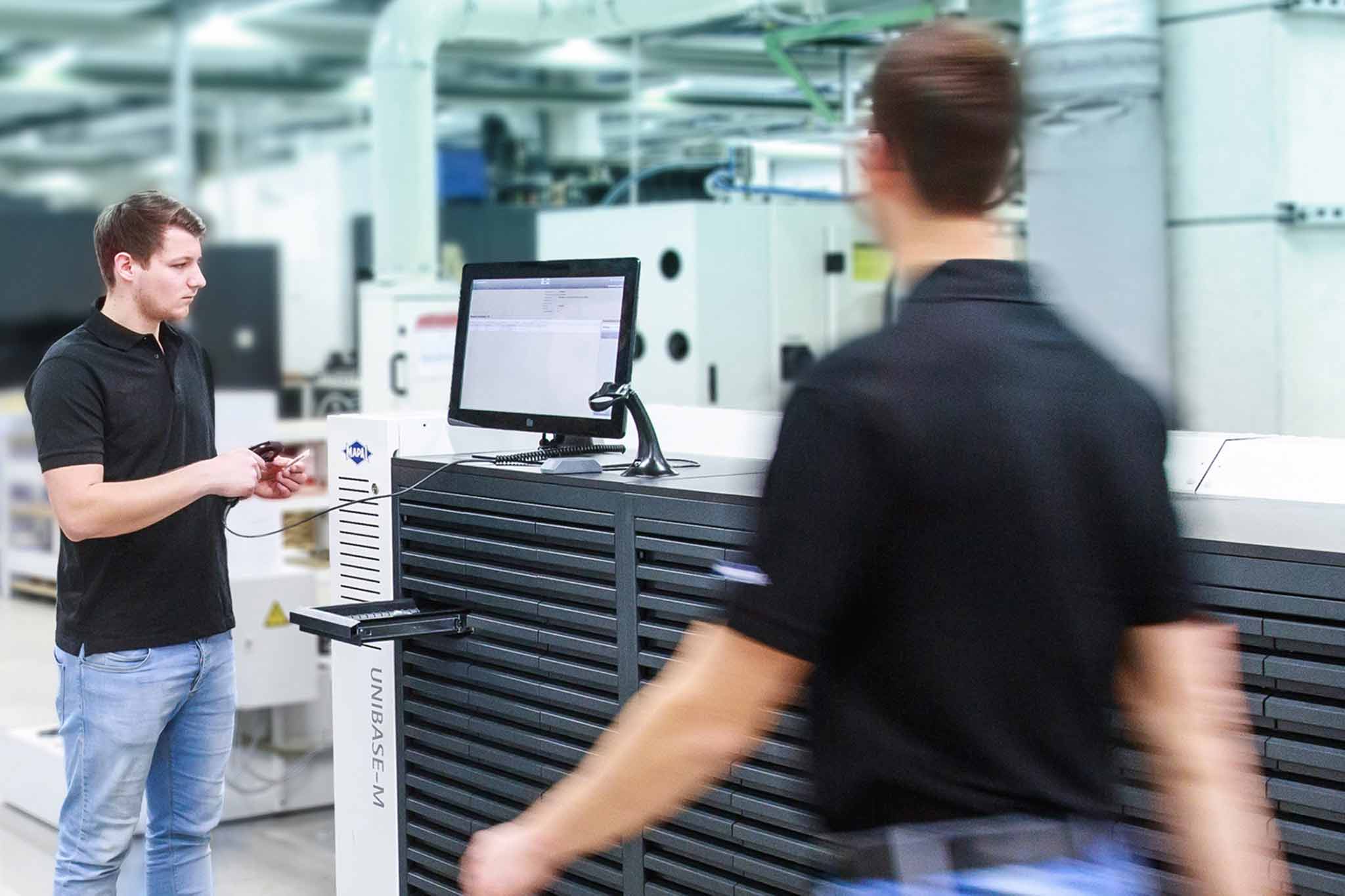 Ein Mitarbeiter steht vor einem Werkzeugausgabesystem UNIBASE-M. Ein Mitarbeiter läuft vorbei. Ein Mitarbeiter steht vor einem Werkzeugausgabesystem UNIBASE-M. Ein Mitarbeiter läuft vorbei.