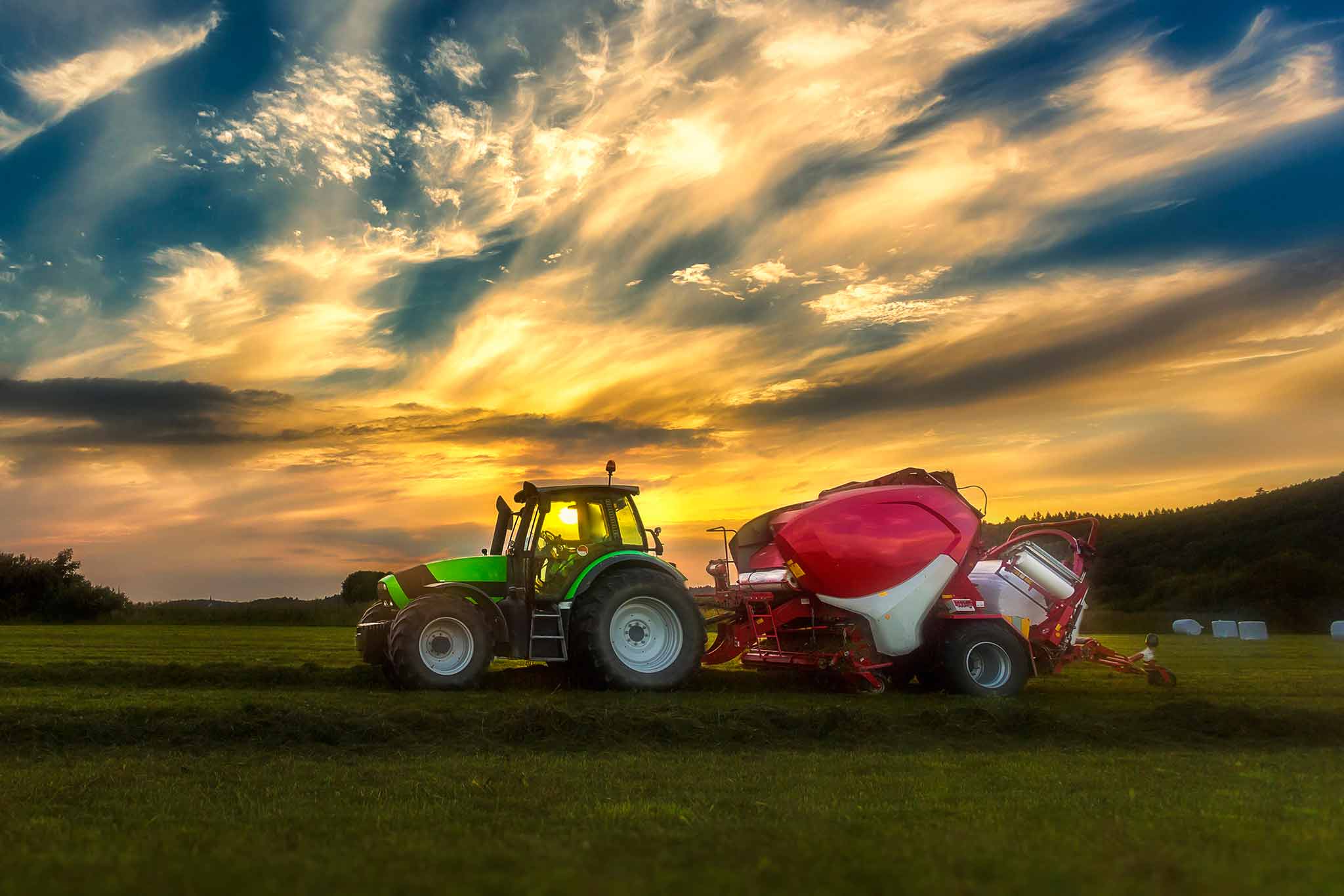 A tractor is shown in a meadow in front of a cloudy sky with a round baler attached. 