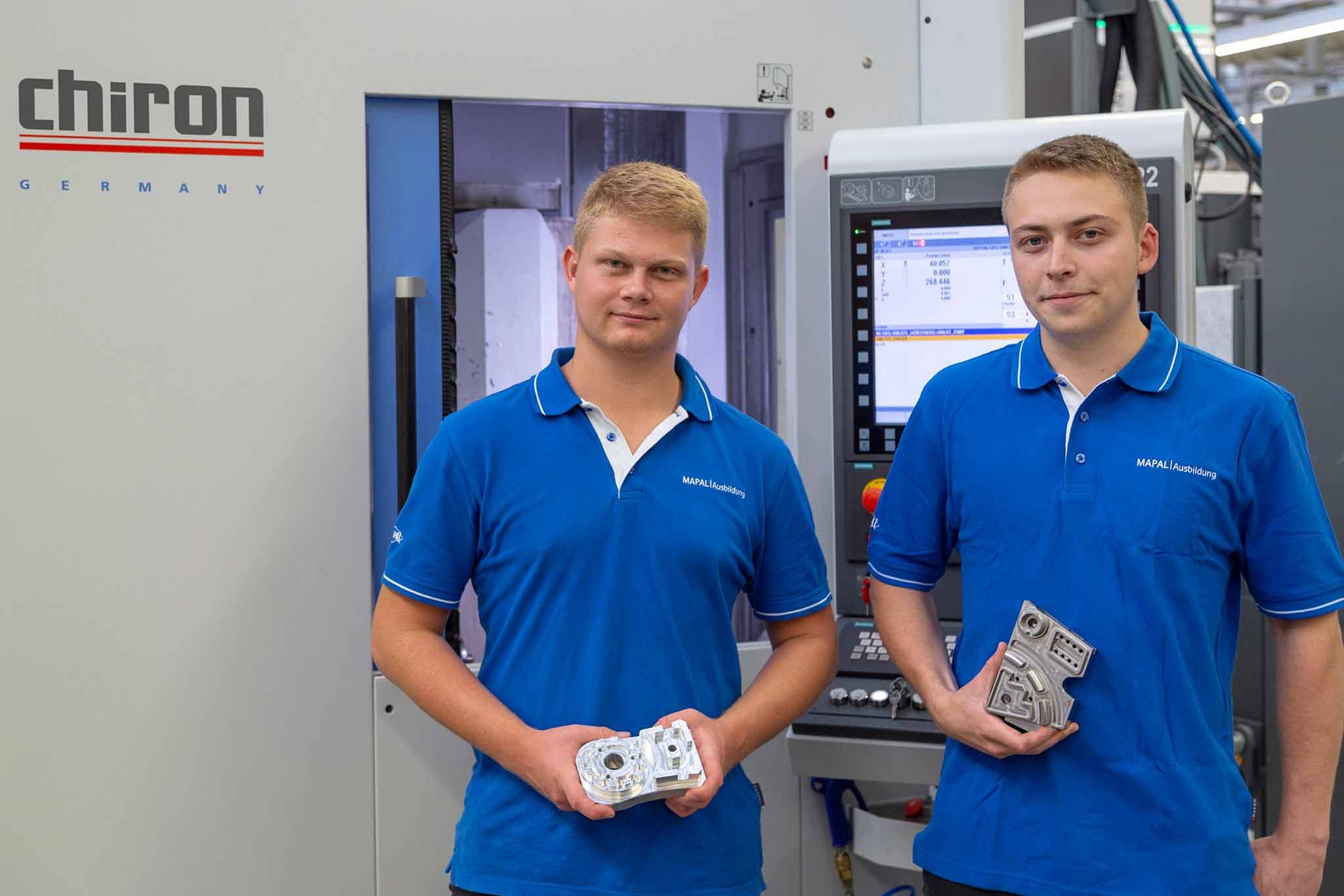 Julian Kraus and Lukas Weiss (from left) in front of a Chiron machine. Both of them are holding a part in their hands.